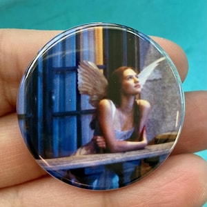 May include: A round button with a picture of a woman with wings looking up at the sky. The woman is wearing a white dress and has long brown hair. The background is a blue and white window.