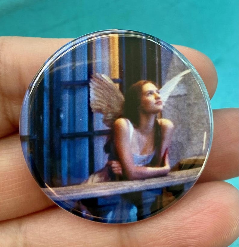 May include: A round button with a picture of a woman with wings looking up at the sky. The woman is wearing a white dress and has long brown hair. The background is a blue and white window.