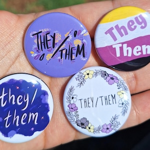 May include: Four round pronoun pins with the text "They/Them". The pins have various designs, including a purple background with floral accents, a non-binary flag design, a blue watercolor effect, and a floral wreath.