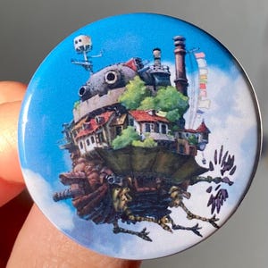 May include: A round button with a blue background featuring a detailed illustration of a flying house from the animated film 'Howl's Moving Castle'. The house is depicted with a chimney, windows, and a green roof. It is surrounded by clouds and has a pair of bird-like legs.