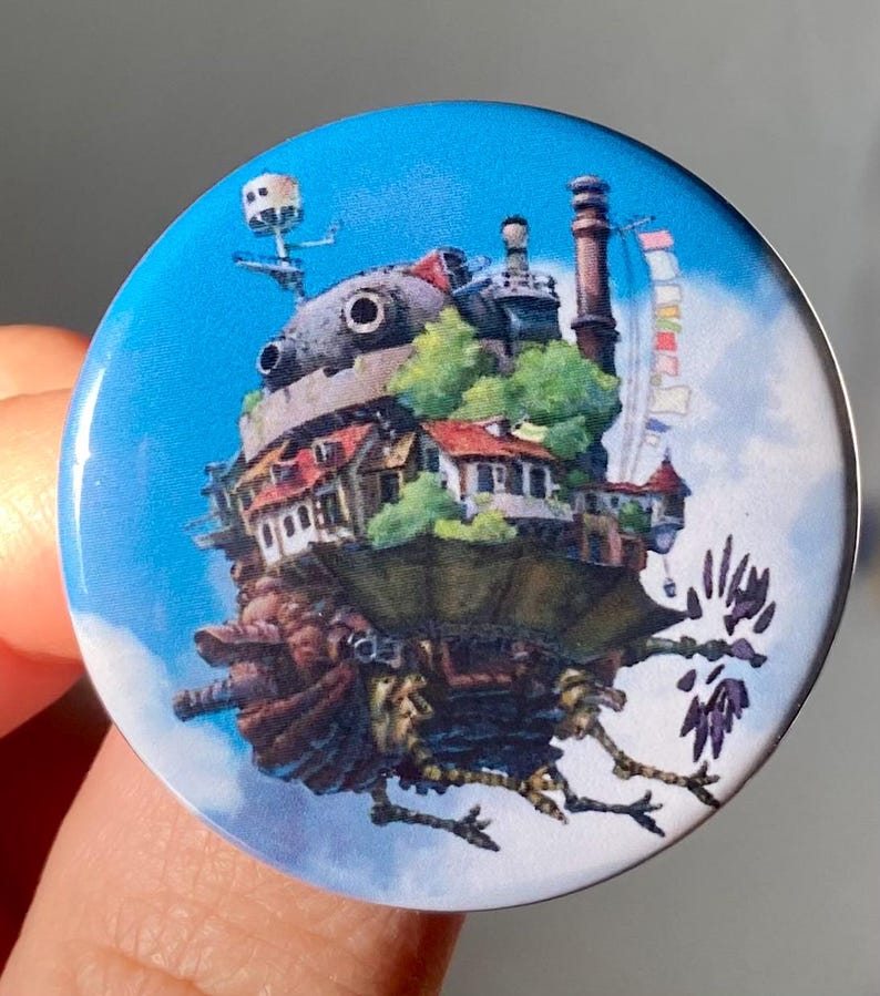 May include: A round button with a blue background featuring a detailed illustration of a flying house from the animated film 'Howl's Moving Castle'. The house is depicted with a chimney, windows, and a green roof. It is surrounded by clouds and has a pair of bird-like legs.