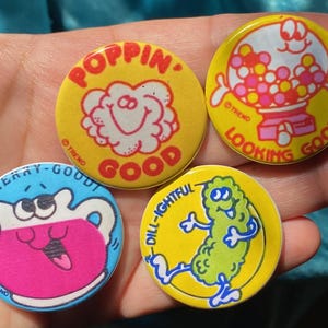 Puede incluir: Cuatro botones redondos amarillos y blancos con personajes de dibujos animados y texto. Los botones dicen "Cherry-Good!" con una jarra de dibujos animados de jugo de cereza, "Poppin' Good!" con una nube de dibujos animados, "Dill-Ightiful" con un pepinillo de dibujos animados y "Looking Good!" con una máquina de chicles de dibujos animados.