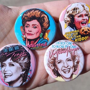 May include: A collection of four round pinback buttons featuring portraits of a woman. Each button has a different background color and text, including "Flirting is part of my heritage," "Sometimes life just isn't fair, kiddo!", "Eat dirt and die trash," and "I'm not one to blow my own Vertuberflugen."