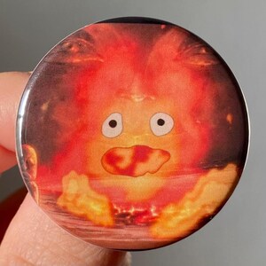 May include: A round button with a cartoon image of a fire spirit from the movie "My Neighbor Totoro". The spirit is orange and red with a large, open mouth and two large, white eyes.