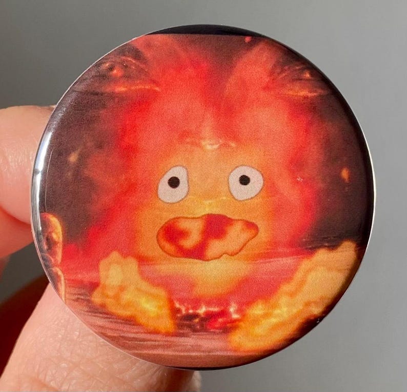 May include: A round button with a cartoon image of a fire spirit from the movie "My Neighbor Totoro". The spirit is orange and red with a large, open mouth and two large, white eyes.