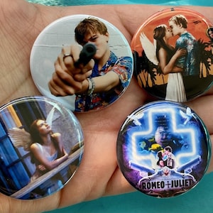 May include: Four round button pins featuring scenes from the movie Romeo + Juliet. The first pin shows Leonardo DiCaprio holding a gun. The second pin shows Claire Danes and Leonardo DiCaprio embracing. The third pin shows Claire Danes standing on a balcony. The fourth pin shows a neon cross with the text "Romeo + Juliet" underneath.
