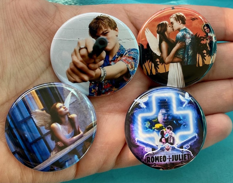 May include: Four round button pins featuring scenes from the movie Romeo + Juliet. The first pin shows Leonardo DiCaprio holding a gun. The second pin shows Claire Danes and Leonardo DiCaprio embracing. The third pin shows Claire Danes standing on a balcony. The fourth pin shows a neon cross with the text "Romeo + Juliet" underneath.