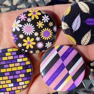 May include: Four round buttons with different designs. The first button has a black background with yellow and purple flowers. The second button has a black background with white and purple leaves. The third button has a black background with yellow and purple squares. The fourth button has a black background with white, yellow, and purple stripes.