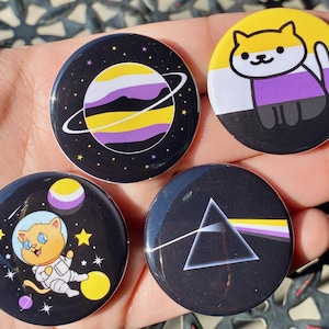 Op de afbeelding: Vier ronde knopen met verschillende ontwerpen. De eerste knoop heeft een gele, witte en paarse planeet met een ring eromheen op een zwarte achtergrond met witte sterren. De tweede knoop heeft een gele, witte en paarse gestreepte achtergrond met een zwarte kat met een wit gezicht en gele ogen. De derde knoop heeft een zwarte kat in een wit ruimtepak met een gele, witte en paarse planeet op de achtergrond. De vierde knoop heeft een zwarte en witte driehoek met een gele, witte en paarse streep aan de onderkant op een zwarte achtergrond.