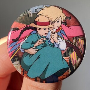 May include: A round button featuring a scene from the animated film Howl's Moving Castle. The button shows Howl and Sophie, with Sophie wearing a straw hat and a teal dress, as they fly through the air.