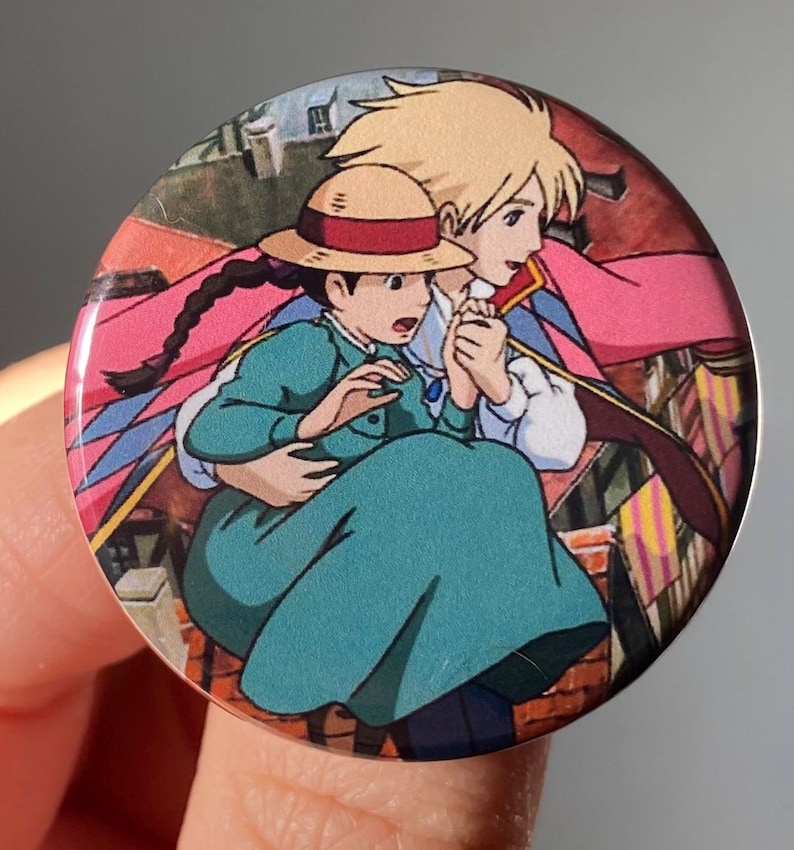 May include: A round button featuring a scene from the animated film Howl's Moving Castle. The button shows Howl and Sophie, with Sophie wearing a straw hat and a teal dress, as they fly through the air.