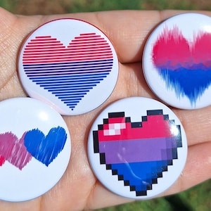 Pode incluir: Quatro botões redondos brancos com desenhos de coração em vermelho, rosa, azul e roxo. Um botão tem um coração pixelizado, outro tem um coração com listras horizontais e um terceiro tem um coração com pinceladas verticais. O quarto botão tem três corações.