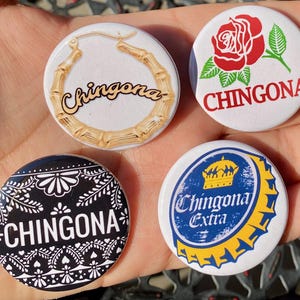 May include: Four round button pins with the word "Chingona" on each. One pin features a gold hoop earring, one features a red rose, one features a black and white bandana pattern, and one features a blue and yellow beer bottle cap.