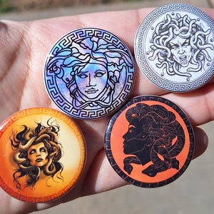 Puede incluir: Cuatro botones redondos con temática de Medusa. Un botón presenta una cara de Medusa azul y morada, otro una cara de Medusa en blanco y negro, y otros dos con fondos naranjas e imágenes de Medusa. Cada botón tiene un borde decorativo.