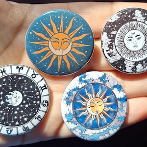 May include: Four circular celestial-themed badges. Designs feature a sun and moon with stars on a blue backdrop, a sun with a face on a monochrome background, a zodiac design, and a sun and moon with clouds on a blue background.