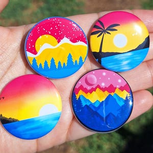 Pode incluir: Quatro botões redondos com designs de paisagens coloridas. Cada botão apresenta um pôr do sol ou uma cena de montanha diferente em tons de rosa, amarelo, azul e branco. Um botão tem uma silhueta de palmeira.
