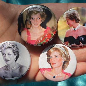 Puede incluir: Cuatro botones redondos con diferentes imágenes de la princesa Diana. Los botones son blancos con un borde plateado. Las imágenes son en color y en blanco y negro.
