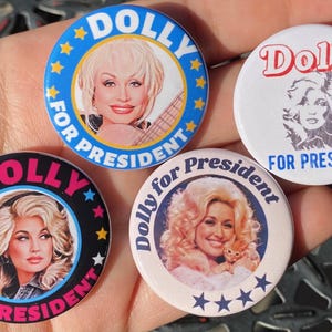 May include: Four round buttons with a black, white, and blue color scheme. Each button features a portrait of Dolly Parton with the text "Dolly for President" in a white, pink, and blue font.