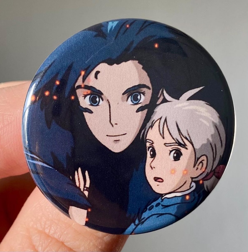 May include: A round button featuring a cartoon image of two characters from the animated film "Howl's Moving Castle". The characters are a young girl with white hair and a man with dark blue hair. The background is a dark blue with orange sparks.