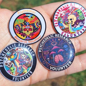 Könnte beinhalten: Vier runde, farbenfrohe Anstecknadeln mit psychedelischen Designs. Jede Anstecknadel trägt die Worte "Psychedelic Research Volunteer" in einem kreisförmigen Muster. Die Designs umfassen Schädel, Außerirdische, Pilze und wirbelnde Muster.