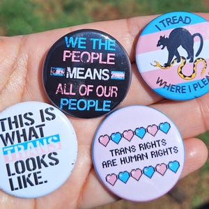 Op de afbeelding: Vier ronde buttons met tekst en afbeeldingen. Eén button zegt "WE THE PEOPLE MEANS ALL OF OUR PEOPLE". Een andere zegt "THIS IS WHAT TRANS LOOKS LIKE". Een derde button zegt "TRANS RIGHTS ARE HUMAN RIGHTS". De laatste button zegt "I TREAD WHERE I PLEASE".