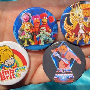 Puede incluir: Cuatro botones redondos con diferentes personajes de dibujos animados. El primer botón presenta un fondo azul con cuatro personajes de los Muppets. El segundo botón presenta un fondo blanco con un arcoíris y el texto "Rainbow Brite". El tercer botón presenta un fondo negro con el logotipo de He-Man y los Masters del Universo. El cuarto botón presenta un fondo azul con el logotipo de She-Ra Princesa del Poder.