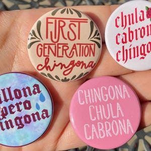 Könnte beinhalten: Vier runde Knöpfe mit verschiedenen Designs und Sätzen. Der erste Knopf ist beige mit einem roten und schwarzen Blumendesign und dem Text "First Generation Chingona". Der zweite Knopf ist weiß mit einer roten Rose und dem Text "Chula Cabrona Chingona". Der dritte Knopf ist blau mit einem weißen und violetten Aquarelldesign und dem Text "Chillona Pero Chingona". Der vierte Knopf ist rosa mit weißem Text, der "Chingona Chula Cabrona" lautet.
