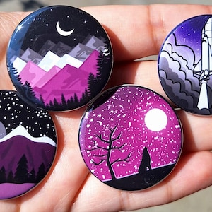 Puede incluir: Cuatro pines redondos con diseños de paisajes. Uno presenta una luna creciente sobre montañas, otro un cohete despegando y otros dos con escenas de noche estrellada. Los pines tienen un acabado brillante y se sostienen en una mano.