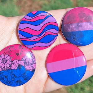 Pode incluir: Quatro botões redondos e brilhantes com designs de orgulho bissexual. Um apresenta linhas onduladas rosa, roxas e azuis. Outro tem um design floral rosa e azul. Os outros dois têm as cores da bandeira bissexual.