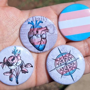 Op de afbeelding: Vier ronde knopen met verschillende ontwerpen. Eén knoop toont de transgender vlag, een andere een illustratie van een menselijk hart, een derde een roos en de laatste een kompasroos. De knopen zijn in blauw, roze en wit.