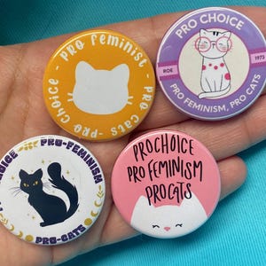Puede incluir: Cuatro botones redondos con diferentes diseños. Cada botón presenta una ilustración de un gato y un texto que dice "Pro-Choice", "Pro-Feminismo", "Pro-Cats" y "Roe 1973".