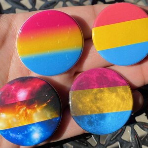 Pode incluir: Quatro botões redondos com um design de bandeira pansexual. Os botões apresentam um esquema de cores rosa, amarelo e azul com um padrão de galáxia em dois dos botões.