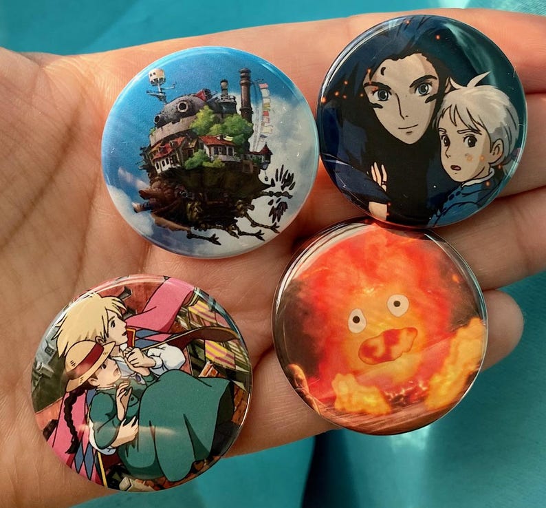 May include: Four round buttons with different images from the animated film 'Howl's Moving Castle'. The first button features a close-up of Howl and Calcifer, the second button features a close-up of the moving castle, the third button features a close-up of the fire demon, and the fourth button features a close-up of Sophie and Howl.