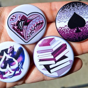 Pode incluir: Quatro botões redondos com designs únicos. Um apresenta um coração com um padrão geométrico, outro uma pá preta sobre um fundo roxo, um terceiro um dragão com uma pá e o último uma pilha de livros. Os botões são brancos com ilustrações coloridas.