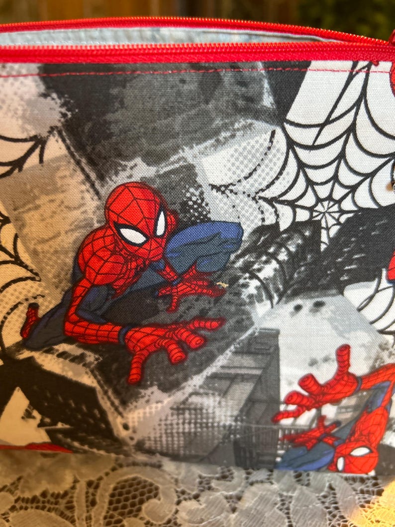 Puede incluir: Una bolsa roja con cremallera con un dise&ntilde;o de Spider-Man. La bolsa tiene una cremallera roja y un fondo negro, blanco y gris con gr&aacute;ficos de Spider-Man y telara&ntilde;as. La bolsa est&aacute; hecha de tela.
