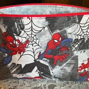 Puede incluir: Una bolsa rectangular con cremallera con un dise&ntilde;o de Spider-Man. La bolsa tiene una cremallera y un ribete rojos, con un fondo negro, blanco y gris. Spider-Man se representa en varias poses de acci&oacute;n, con telara&ntilde;as.