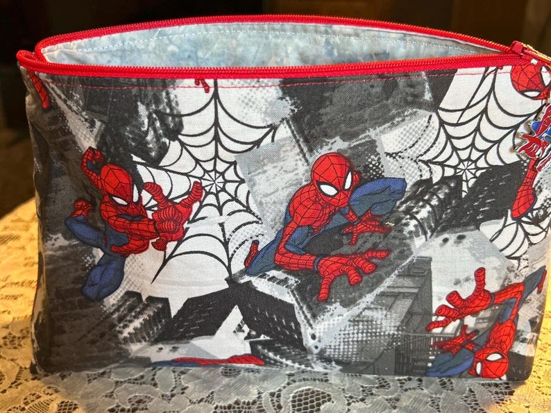 Puede incluir: Una bolsa rectangular con cremallera con un dise&ntilde;o de Spider-Man. La bolsa tiene una cremallera y un ribete rojos, con un fondo negro, blanco y gris. Spider-Man se representa en varias poses de acci&oacute;n, con telara&ntilde;as.