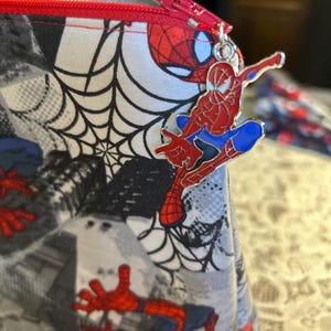 Puede incluir: Una bolsa con cremallera con tem&aacute;tica de Spider-Man. La tela muestra un patr&oacute;n de Spider-Man, telara&ntilde;as y paisajes urbanos en rojo, negro, blanco y azul. Una cremallera roja y un dije de Spider-Man completan el dise&ntilde;o.