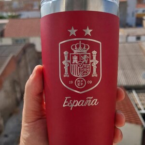 May include: A red stainless steel tumbler featuring a white laser-engraved Spanish national football team crest with two stars above it and the word España written below. The cup has a clear plastic lid and a metallic silver rim at the top and base.