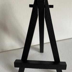 May include: A small, black wooden easel with a three-legged design. The easel has a rectangular base and a vertical support structure, ideal for displaying artwork or photos. The wood has a matte finish.