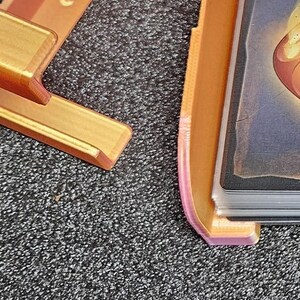 May include: Three 3D printed card holders, one orange, one pink, and one black. Each holder has a slot for a card and a stand to hold it upright. The orange and pink holders have a card with a graphic design in them, while the black holder has a card with a comic book character on it.