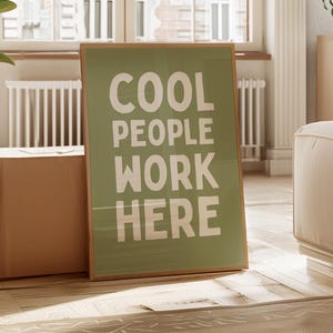 Puede incluir: Un póster enmarcado con el texto "COOL PEOPLE WORK HERE" en blanco sobre un fondo verde. El póster está apoyado contra una caja marrón en una habitación luminosa con una ventana grande y una planta.
