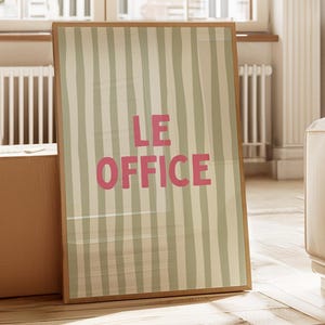 Peut inclure: Une affiche encadrée rectangulaire présentant un motif à rayures verticales dans des tons beige et gris clair. Le texte LE OFFICE est imprimé au centre dans une police rose grasse sans empattement. Le cadre est en bois clair.
