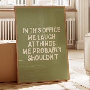 May include: A framed poster with the text "IN THIS OFFICE WE LAUGH AT THINGS WE PROBABLY SHOULDN'T" in white letters on a green background. The frame is a light brown color.