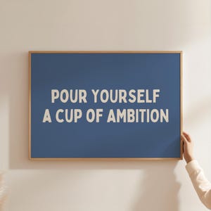 Può includere: Una stampa da parete rettangolare in una cornice di legno chiaro con uno sfondo blu solido e il testo "POUR YOURSELF A CUP OF AMBITION" stampato in un carattere sans-serif grassetto color bianco sporco centrato al centro della stampa.