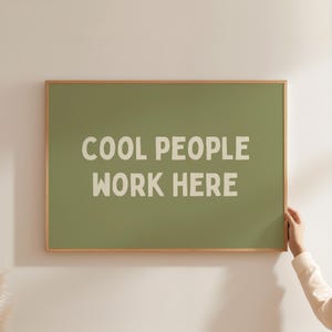 Op de afbeelding: Een rechthoekig, ingelijst bord met de tekst "COOL PEOPLE WORK HERE" in witte letters op een groene achtergrond. De lijst is van lichtbruin hout. Een hand houdt het frame vast.