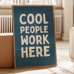 Pode incluir: Uma peça de arte retangular emoldurada com um fundo azul escuro e o texto COOL PEOPLE WORK HERE impresso em letras maiúsculas grandes e arrojadas na cor branco-sujo. A moldura é feita de madeira clara com um design simples.