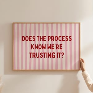 May include: A rectangular wall art piece in a light wooden frame featuring a background of vertical pink and white stripes. The text "DOES THE PROCESS KNOW WE'RE TRUSTING IT?" is printed in bold, dark red, sans-serif capital letters in the centre.