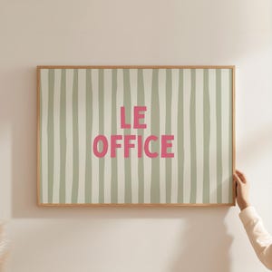 Puede incluir: Un cuadro enmarcado con el texto "LE OFFICE" en letras rosas. El fondo presenta rayas verticales en verde claro y blanco. El marco es de madera marrón claro.