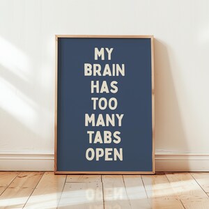May include: A framed print with the text "MY BRAIN HAS TOO MANY TABS OPEN" in white letters on a dark blue background. The frame is light brown, and the print is against a white wall and wooden floor.